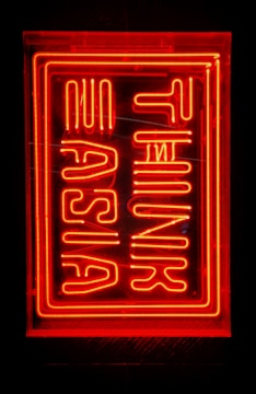 Bright red neon sign with stylized lettering, possibly spelling out 'Turn Up Asia'. The sign is enclosed in a rectangular frame against a dark background.