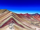 A vibrant mountainous landscape with layers of red, yellow, and brown hues creating a striped effect. A trail of people walking along a path adds a sense of scale. The sky is clear, with a deep blue tone contrasting the earthy colors.