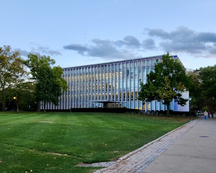 A modern, multi-story building with glass panels is surrounded by lush green trees and a well-maintained lawn. The sky above is partly cloudy, suggesting a calm, overcast day.