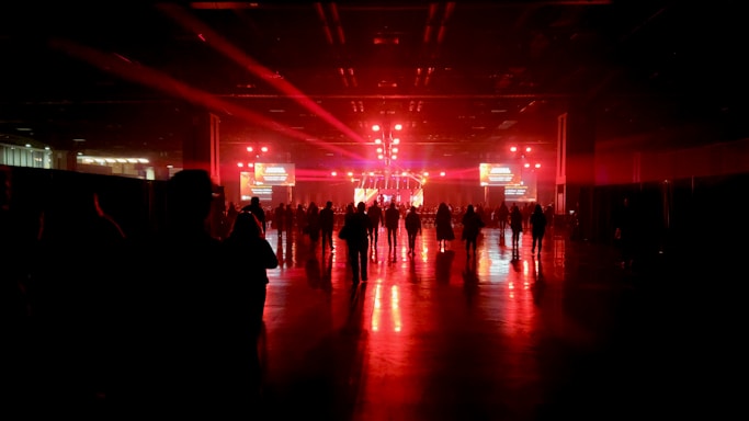 A large, dimly lit indoor venue with red ambient lighting. A crowd of people stands in front of a stage equipped with bright lights, screens, and speakers. The shiny floor reflects the vivid red coloring, creating a dramatic atmosphere.