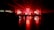 A large, dimly lit indoor venue with red ambient lighting. A crowd of people stands in front of a stage equipped with bright lights, screens, and speakers. The shiny floor reflects the vivid red coloring, creating a dramatic atmosphere.