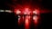 A large, dimly lit indoor venue with red ambient lighting. A crowd of people stands in front of a stage equipped with bright lights, screens, and speakers. The shiny floor reflects the vivid red coloring, creating a dramatic atmosphere.
