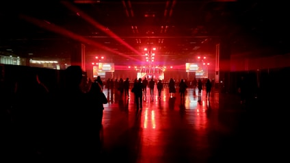 A large, dimly lit indoor venue with red ambient lighting. A crowd of people stands in front of a stage equipped with bright lights, screens, and speakers. The shiny floor reflects the vivid red coloring, creating a dramatic atmosphere.