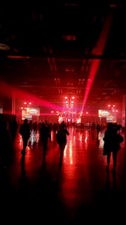 A dimly lit indoor environment with silhouettes of people walking towards a well-lit stage. The stage is illuminated with vibrant red lighting and spotlights, reflecting off the shiny floor, giving the space a dramatic atmosphere. There are large screens on either side of the stage displaying text or images.