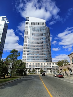 A tall, modern high-rise building stands against a backdrop of a bright blue sky scattered with fluffy white clouds. The building features a sleek, curved design with numerous windows reflecting the sky. In the foreground, a paved road leads directly to the complex, bordered by a few trees and parked cars.