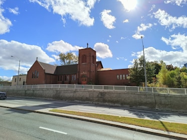 A red brick church with a tall bell tower is situated along a street. The building is surrounded by a white fence and lush greenery, with a clear blue sky and fluffy white clouds above. The sun is shining brightly, casting shadows on the road and the church.