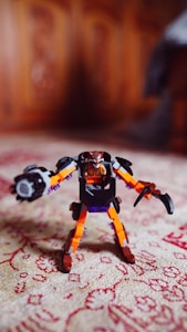 A detailed view of a colorful toy robot made of plastic blocks standing on a patterned carpet. The robot has orange, black, and purple components, with distinct robotic arms and a humanoid head. The background is softly blurred, focusing attention on the toy.