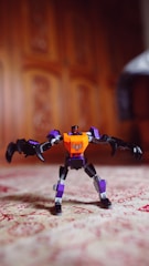 A toy robot with an orange and purple body stands on a patterned surface. The background features a wooden texture with intricate designs.