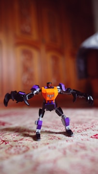 A toy robot with an orange and purple body stands on a patterned surface. The background features a wooden texture with intricate designs.