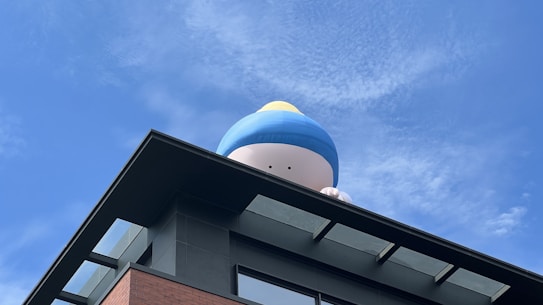 A large inflatable figure resembling a cartoon character is positioned on the roof of a building, partially visible with just the top portion of its head and hands showing. The character wears a blue and yellow hat. The building has a modern architectural style with black edges and a mix of black and red brick surfaces. The sky is clear with some patches of white clouds.