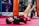 Children are engaging in a martial arts or fitness class, lying on their backs with legs raised. They are in a gym setting with red mats and practice bags visible in the background.