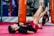 Children are engaging in a martial arts or fitness class, lying on their backs with legs raised. They are in a gym setting with red mats and practice bags visible in the background.