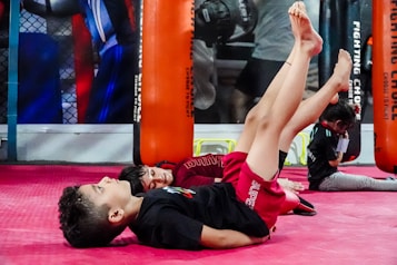Children are engaging in a martial arts or fitness class, lying on their backs with legs raised. They are in a gym setting with red mats and practice bags visible in the background.