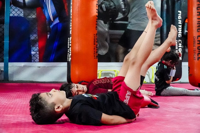 Children are engaging in a martial arts or fitness class, lying on their backs with legs raised. They are in a gym setting with red mats and practice bags visible in the background.