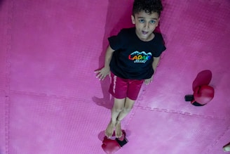 A young child with curly hair is lying on a pink exercise mat. The child is wearing a black t-shirt with a colorful design and red shorts. Two red boxing gloves are placed on the mat, one near the child's head and the other near their feet.