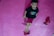 A young child with curly hair is lying on a pink exercise mat. The child is wearing a black t-shirt with a colorful design and red shorts. Two red boxing gloves are placed on the mat, one near the child's head and the other near their feet.