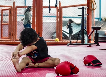 A young child stretches on a red mat in a martial arts or gym setting, surrounded by protective fencing. The child is focused, wearing a black shirt and red shorts, with red boxing gloves placed nearby. In the background, two other people can be seen, one walking and the other standing, suggesting a gymnasium environment.