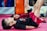 Two children are lying on their backs on a pink mat, performing exercises with their legs raised. One child is wearing a black shirt with colorful graphics, while the other wears a dark red shirt. An orange vertical padded structure is visible in the background.
