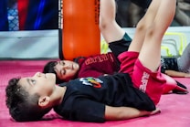 Two children are lying on their backs on a pink mat, performing exercises with their legs raised. One child is wearing a black shirt with colorful graphics, while the other wears a dark red shirt. An orange vertical padded structure is visible in the background.