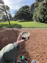 A person holding a cup of mate in a park setting with a thermos and shoes visible. The ground is covered in red gravel, and the background features lush green grass and trees on a sunny day, with a lamp post visible on the left.