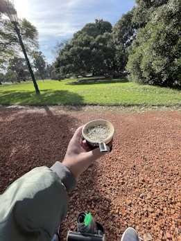 A person holding a cup of mate in a park setting with a thermos and shoes visible. The ground is covered in red gravel, and the background features lush green grass and trees on a sunny day, with a lamp post visible on the left.