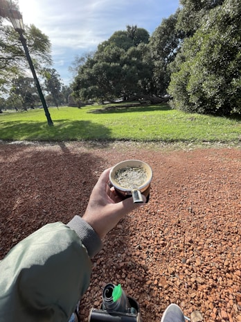 A person holding a cup of mate in a park setting with a thermos and shoes visible. The ground is covered in red gravel, and the background features lush green grass and trees on a sunny day, with a lamp post visible on the left.