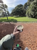 A person holding a cup of mate in a park setting with a thermos and shoes visible. The ground is covered in red gravel, and the background features lush green grass and trees on a sunny day, with a lamp post visible on the left.