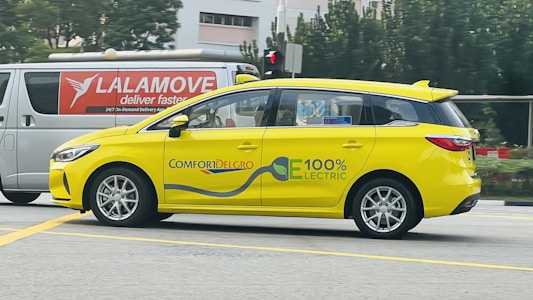A bright yellow electric car with the words 'ComfortDelGro' and '100% Electric' on its side is driving on a road. Its modern design is complemented by a sleek body and silver rims. In the background, a white van with red lettering advertising delivery services is visible against a backdrop of green trees and an urban setting.
