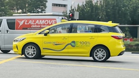 A bright yellow electric car with the words 'ComfortDelGro' and '100% Electric' on its side is driving on a road. Its modern design is complemented by a sleek body and silver rims. In the background, a white van with red lettering advertising delivery services is visible against a backdrop of green trees and an urban setting.