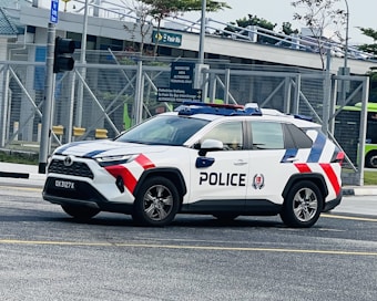 A police SUV with a distinctive white, red, and blue color scheme is parked on a road near a sign indicating 'Pasir Ris'. The vehicle has a prominent police emblem and text on the side. In the background, there's a metal fence, a building, a traffic light, and some greenery.