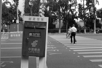 A black and white scene showing a China Post mailbox at the forefront with a street intersection in the background. A person is seen crossing the crosswalk with a bicycle under a canopy of trees.