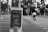 A black and white scene showing a China Post mailbox at the forefront with a street intersection in the background. A person is seen crossing the crosswalk with a bicycle under a canopy of trees.