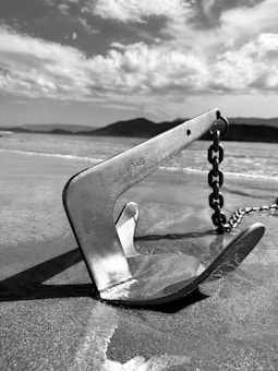 A metallic anchor rests on sandy ground near the water's edge, with a chain attached. The background features a calm sea with gentle waves and a distant view of mountains under a sky populated with soft, scattered clouds.
