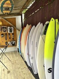 A selection of surfboards is neatly lined up against a wall with a variety of colors, including white, yellow, and orange. Above, a rack displays various surfing accessories, such as leashes and fins. The floor is wooden, and a folding table stands close to the wall. A decorative surfboard sign hangs overhead, contributing to the overall beach-themed atmosphere.