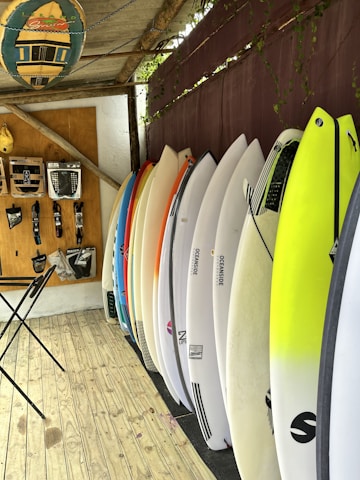 A selection of surfboards is neatly lined up against a wall with a variety of colors, including white, yellow, and orange. Above, a rack displays various surfing accessories, such as leashes and fins. The floor is wooden, and a folding table stands close to the wall. A decorative surfboard sign hangs overhead, contributing to the overall beach-themed atmosphere.