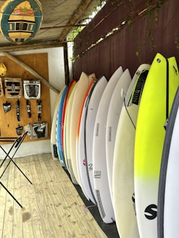 A selection of surfboards is neatly lined up against a wall with a variety of colors, including white, yellow, and orange. Above, a rack displays various surfing accessories, such as leashes and fins. The floor is wooden, and a folding table stands close to the wall. A decorative surfboard sign hangs overhead, contributing to the overall beach-themed atmosphere.