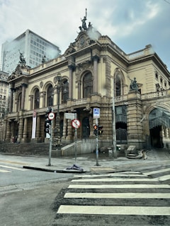 A grand historic theater building with intricate architectural details and classical statues on the facade. The structure features large columns and arched windows, with a modern high-rise building visible in the background. The street in the foreground is wet, possibly from rain, and there are several traffic signs and lights.