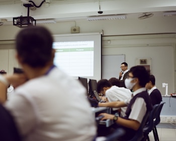 A classroom or conference room setting with a group of people seated at desks, looking towards a speaker and a projected slideshow. The speaker stands at the front near a projection screen displaying a table. Most attendees are wearing formal attire and some have masks on.