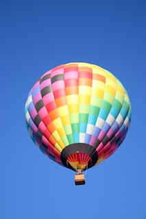 A vibrant, multicolored hot air balloon floats against a clear blue sky. The balloon's panels are arranged in a pattern of alternating bright colors including red, orange, yellow, green, blue, and purple. The wicker basket is visible below, hanging from ropes attached to the balloon.