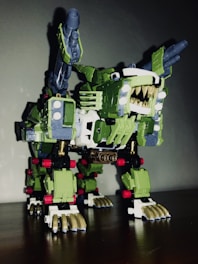 A robotic toy resembling a mechanical creature with sharp teeth, claws, and missile launchers on its back. It has a predominantly green body with white and gold detailing and some red accents. The design is intricate, featuring various mechanical components and joints.
