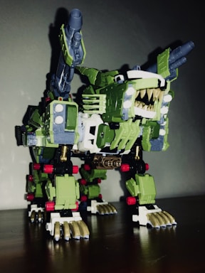 A robotic toy resembling a mechanical creature with sharp teeth, claws, and missile launchers on its back. It has a predominantly green body with white and gold detailing and some red accents. The design is intricate, featuring various mechanical components and joints.