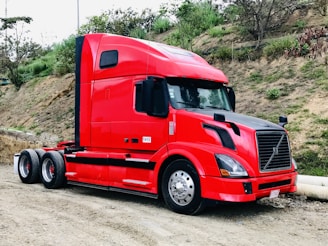 A bright red semi-truck is parked on a dirt road next to a grassy embankment with some scattered bushes and trees. The truck is a modern design with a streamlined cab and large side mirrors. It stands out against the natural background.
