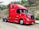 A bright red semi-truck is parked on a dirt road next to a grassy embankment with some scattered bushes and trees. The truck is a modern design with a streamlined cab and large side mirrors. It stands out against the natural background.