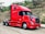 A bright red semi-truck is parked on a dirt road next to a grassy embankment with some scattered bushes and trees. The truck is a modern design with a streamlined cab and large side mirrors. It stands out against the natural background.