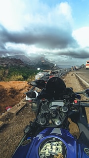 A motorcycle is parked on the side of a scenic mountain road with a view of the landscape. The sky is overcast, with dark, moody clouds hanging low, casting a dramatic tone over the surroundings. In the background, another motorcycle is visible, along with a few vehicles on the road.