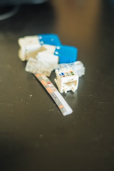 A collection of small, disassembled plastic electronic components rests on a dark surface. A blue plastic piece and a small, labeled strip are visible among the components.