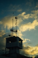 A silhouetted watchtower topped with antennas stands against a dramatic sky filled with clouds that are tinged with the colors of sunset. Barbed wire is visible along the perimeter, suggesting security or surveillance.