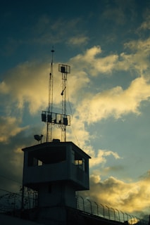 A silhouetted watchtower topped with antennas stands against a dramatic sky filled with clouds that are tinged with the colors of sunset. Barbed wire is visible along the perimeter, suggesting security or surveillance.