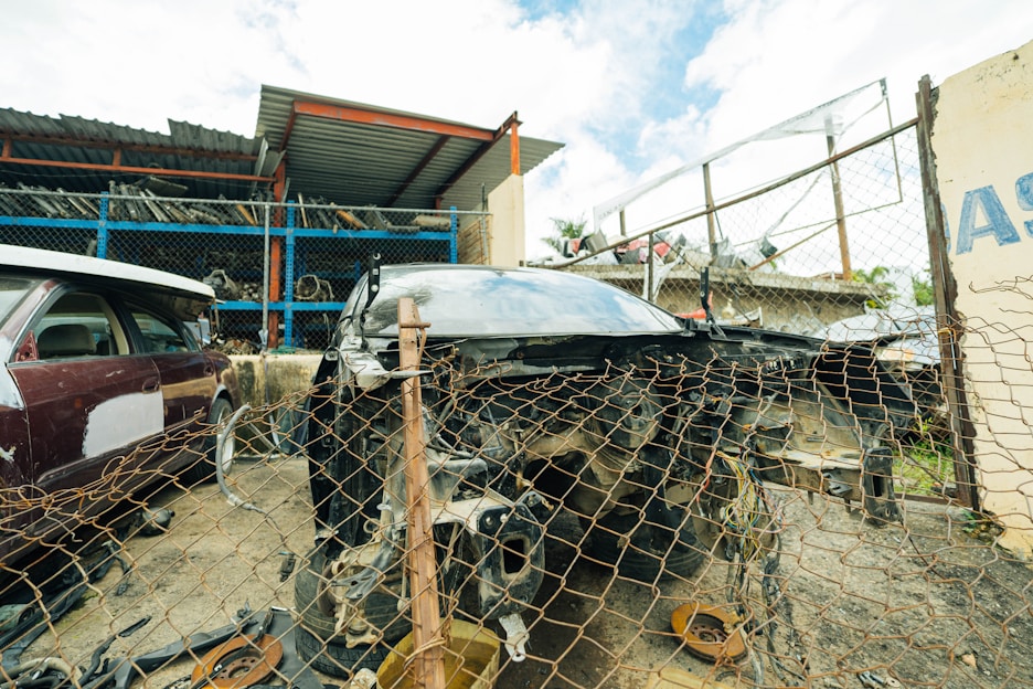 A damaged car is positioned behind a rusty chain-link fence in a scrapyard setting. Surrounding the car are various automotive parts scattered on the ground. In the background, partially visible vehicles and metal shelving filled with disassembled car components can be seen. The area is enclosed by a metal fence, and a small structure with a corrugated metal roof is nearby.