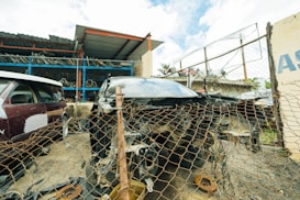 A damaged car is positioned behind a rusty chain-link fence in a scrapyard setting. Surrounding the car are various automotive parts scattered on the ground. In the background, partially visible vehicles and metal shelving filled with disassembled car components can be seen. The area is enclosed by a metal fence, and a small structure with a corrugated metal roof is nearby.